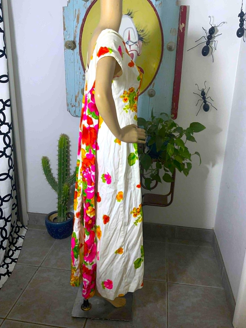 May include: White vintage maxi dress with a vibrant floral print in shades of pink, orange, and yellow. The dress features short sleeves and a flowing silhouette. The floral pattern is concentrated on the side and scattered across the front.