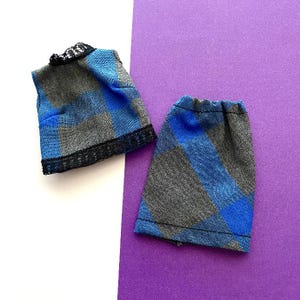 May include: A doll-sized blue, gray, and black plaid vest and skirt set. The vest has a black lace trim around the neckline and bottom. The skirt is a matching plaid pattern with a gathered waist. The set is displayed on a white and purple background.