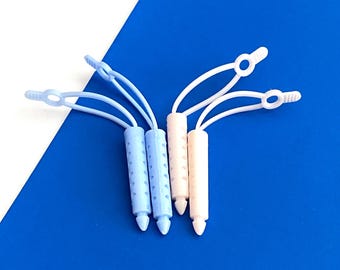 May include: Four plastic zipper pulls with flexible loops. Two pulls are light blue and two are off-white. Each pull features a cylindrical body with a textured grip surface and a pointed tip, attached to a thin, looped cord with a flat end tab.