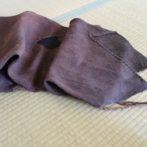 Puede incluir: Una pieza textil rectangular de color marrón oscuro con una textura tejida gruesa. La tela presenta un pequeño parche oscuro en forma de hoja cosido en la superficie e incluye un cordón fino y deshilachado que sobresale de una esquina.