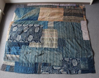 May include: A patchwork quilt with a variety of blue, tan, and white fabrics. The quilt features floral and striped patterns, with a mix of textures and shades. The quilt is rectangular and appears to be handmade.