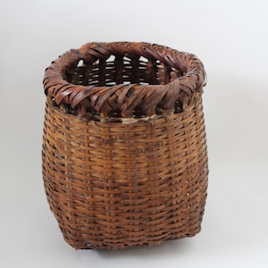 Könnte beinhalten: Ein handgefertigter, zylindrischer Korb aus natürlichen braunen Schilfrohren. Der Korb hat einen dicht gewebten Körper und einen dekorativen, geflochtenen Rand. Die offene Oberseite und die robuste Konstruktion deuten darauf hin, dass er für die Aufbewahrung oder Präsentation bestimmt ist.