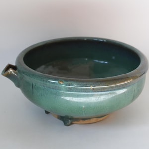 Puede incluir: Un cuenco de cerámica turquesa con un pico y tres patas pequeñas. El cuenco tiene forma redondeada con un borde oscuro y una base de color claro. El esmalte tiene un acabado brillante.