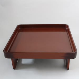 Op de afbeelding: Een vierkant houten dienblad met een rijke, roodbruine afwerking. De lade heeft een opstaande rand en wordt ondersteund door vier korte poten. Het ontwerp is eenvoudig en elegant, geschikt voor het serveren van eten of het uitstallen van items. De afmetingen van de lade zijn ongeveer 30 cm vierkant.