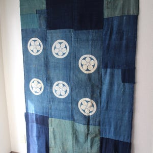 Pode incluir: Um grande tecido retangular com um design de patchwork. O tecido é principalmente azul índigo, com seções azul claro e verde-azulado. Motivos florais brancos são espaçados uniformemente no centro. O tecido está pendurado em uma parede.