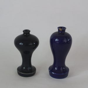 May include: Two small ceramic vases with a glossy finish. One vase is black with a rounded, bulbous body and a narrow neck. The second vase is deep blue with a taller, elongated silhouette and a flared base. Both have a small circular opening.