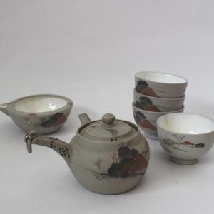 Op de afbeelding: Een keramische theeset met een theepot, een serveerschaal en vijf theekopjes. De set is voornamelijk grijs met handgeschilderde landschapsontwerpen in bruin en zwart. De theekopjes en de kom hebben witte interieurs.