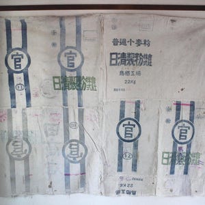 May include: A worn, off-white fabric sack with blue vertical stripes and Japanese text. The sack displays circular seals with Japanese characters and the text "22Kg". The fabric shows signs of age and wear.