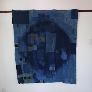 Puede incluir: Un gran tapiz rectangular compuesto por varios parches de tela en tonos azules. La pieza presenta un degradado circular de color índigo oscuro en el centro, rodeado por un mosaico de retazos rectangulares de tela vaquera y algodón.