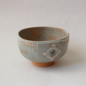 May include: A grey ceramic bowl with a rounded shape and a small, circular base. The bowl features white decorative elements and a central circular design with a Japanese character. The interior has orange speckles.