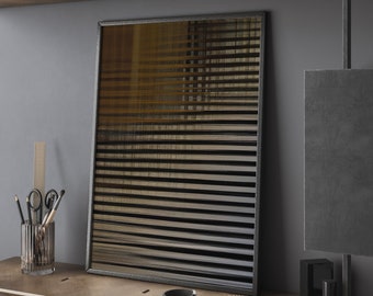 Op de afbeelding: Een ingelijst abstract kunstwerk met horizontale zwarte en gouden lijnen. Het kunstwerk staat op een houten plank met een pennenhouder, schaar en andere decoratieve items. De lijst is donkergrijs.