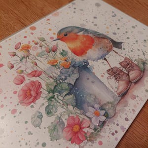 May include: A glass chopping board with a watercolour design of a robin, flowers, and a watering can. The robin has a red breast and grey wings. The flowers are pink, orange, and white. The background has a speckled effect.