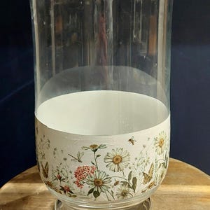 May include: A clear glass vase with a flared top and a round base. The lower section is decorated with a floral pattern of daisies, butterflies, and bees. The vase is on a wooden surface. The height is approximately 30 cm.