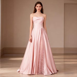 Könnte beinhalten: Ein trägerloses, bodenlanges rosa Kleid. Das Kleid hat ein plissiertes Oberteil und einen fließenden Rock. Ein durchsichtiges Panel mit Perlenakzenten ist im vorderen Schlitz sichtbar. Das Kleid ist aus satinartigem Material.