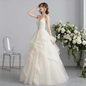 May include: A cream-colored wedding dress with a V-neck bodice and spaghetti straps. The skirt features multiple layers of flowing fabric. A clear chair is in the background, and white flowers are to the right.