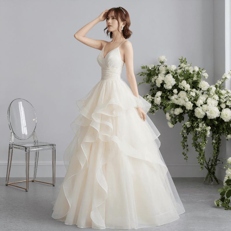 May include: A cream-colored wedding dress with a V-neck bodice and spaghetti straps. The skirt features multiple layers of flowing fabric. A clear chair is in the background, and white flowers are to the right.