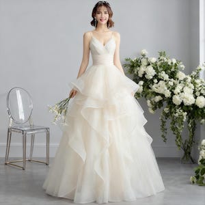May include: A cream-colored wedding dress with a V-neckline and spaghetti straps. The bodice is fitted, and the skirt features layered ruffles. The bride is holding a small bouquet of white flowers. A clear chair is to the left.