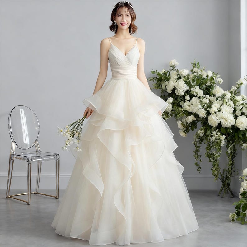 May include: A cream-colored wedding dress with a V-neckline and spaghetti straps. The bodice is fitted, and the skirt features layered ruffles. The bride is holding a small bouquet of white flowers. A clear chair is to the left.