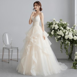 May include: A cream-colored wedding dress with a V-neck bodice and spaghetti straps. The dress features a layered, ruffled skirt and a fitted waist. A clear chair is to the left of the dress, and a bouquet of white flowers is to the right.