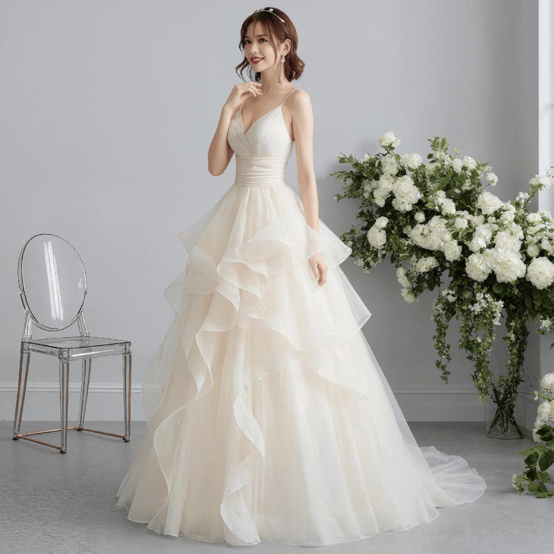May include: A cream-colored wedding dress with a V-neck bodice and spaghetti straps. The dress features a layered, ruffled skirt and a fitted waist. A clear chair is to the left of the dress, and a bouquet of white flowers is to the right.