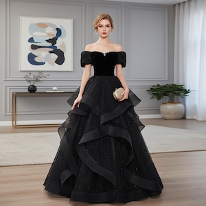 Op de afbeelding: Een zwarte avondjurk met een off-the-shoulder halslijn en een gelaagde rok. De bodice is van fluweel en de rok van tule met subtiele glitters. Een kleine, gouden clutch wordt vastgehouden.