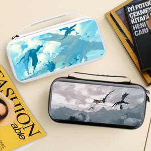 Könnte beinhalten: Zwei rechteckige Etuis mit Drachenillustrationen. Eines ist weiß mit einem blauen Drachendesign vor einem bewölkten Himmel. Das andere ist schwarz mit einer Drachenszene über einer Bergkette. Beide haben einen weißen Griff.