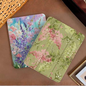 Könnte beinhalten: Zwei Tablet-Hüllen mit floralen Designs. Eine Hülle zeigt ein Design im Aquarellstil mit rosa, lila und blauen Blumen. Die andere Hülle hat einen grünen Hintergrund mit rosa und weißen Blumenmustern.