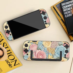 May include: A handheld video game console and a separate console dock, both featuring a floral skin with pink, yellow, and blue roses and hydrangeas. The console has a black screen and black buttons, while the dock displays the full floral pattern.