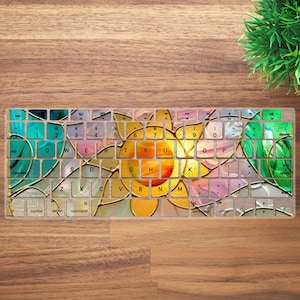 May include: A keyboard cover with a colorful floral design. The design features a large yellow and orange flower in the center, surrounded by green, blue, and pink accents. The keys are visible through the design.