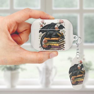 May include: White AirPod case and matching keychain featuring a black dragon resting on a stack of books. The dragon has gold horns and is surrounded by pink flowers and green leaves. The keychain is attached to the case with a silver ring.