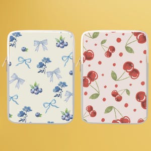 Op de afbeelding: Twee laptophulzen met verschillende patronen. Een hoes heeft een witte achtergrond met blauwe strikken, blauwe bessen en bloemen. De andere hoes heeft een roze achtergrond met rode kersen en groene bladeren.