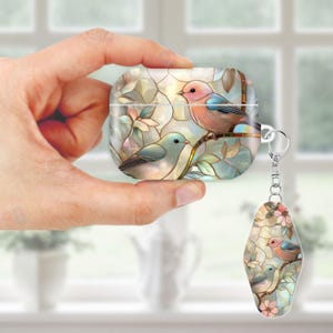 May include: A white AirPod case with a stained glass design featuring two birds perched on a branch with leaves and flowers. A matching keychain with the same design is attached. The case is held in a hand.