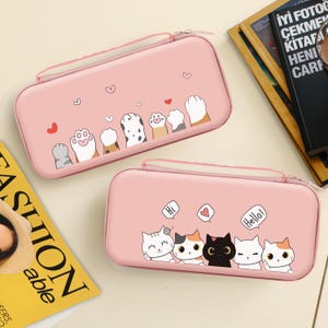 May include: Two pink zippered cases with cat paw prints and hearts. The top case has a row of cat paws. The bottom case features five cartoon cats with speech bubbles that say "Hi" and "Hello".