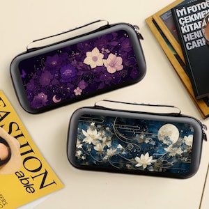 May include: Two black rectangular cases with floral designs. One case features purple flowers, the other has white flowers and a full moon. Both cases have a white handle and a zipper closure. The cases are on a light-colored surface.