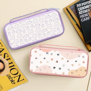 May include: Two rectangular pencil cases. One is lavender with a white background and a pattern of cat faces. The other is pink with a paw print pattern. Both have a zipper closure and a handle.