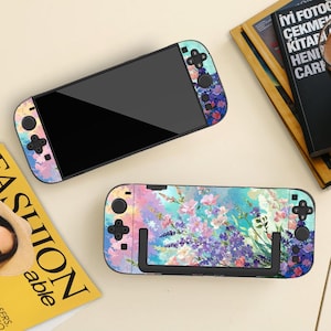 May include: Two Nintendo Switch consoles with floral designs. One console is displayed with the screen facing up, and the other is in a docking station. The floral design features lavender, pink, and blue flowers.