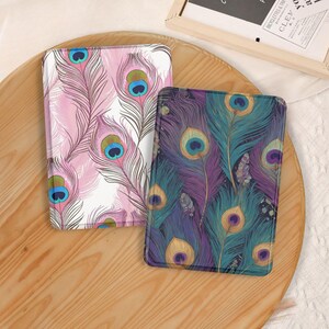 May include: Two rectangular e-reader cases featuring detailed peacock feather patterns. The left case displays pink and white feathers with blue and green eyes. The right case features dark purple and teal feathers with gold and blue eyes.