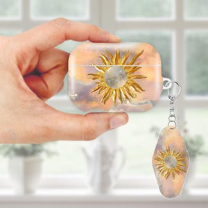 May include: A clear case for wireless earbuds and a matching keychain, both featuring a golden sun and moon design against a watercolor sky of orange, purple, and white. The case is held in a hand.
