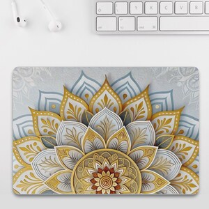 May include: A decorative mousepad featuring a detailed mandala design. The design incorporates gold, white, and blue elements, creating a layered, three-dimensional effect. The mousepad is rectangular and sits beneath a white keyboard.