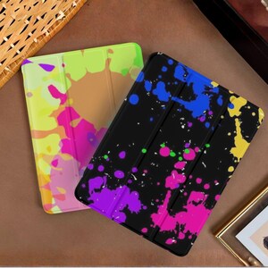 May include: Two rectangular tablet cases featuring vibrant paint splatter designs. One case has a light background with yellow, pink, and orange splashes. The other case has a black background with blue, yellow, purple, and pink paint splatters.