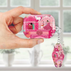 May include: Pink AirPod case and matching keychain featuring a vibrant Indian-inspired design. The case and keychain showcase a detailed illustration of a palace scene with figures, animals, and architectural elements. The design is predominantly pink with accents of other colors.
