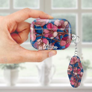 May include: A blue and pink floral design adorns an AirPod case and matching keychain. The case and keychain feature a stained-glass effect with pink blossoms and the name "Oliver" in white script. The keychain is attached to the case with a silver ring.