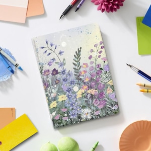 Puede incluir: Un diario con temática floral con un diseño de acuarela que presenta flores moradas, amarillas y blancas. El diario está rodeado de bolígrafos, crayones y muestras de pintura sobre una superficie blanca. El arte de la portada tiene una estética suave y soñadora.