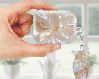 Może przedstawiać: Perłowo-białe etui na AirPods i pasujący brelok do kluczy. Etui i brelok mają płynny, przypominający tkaninę wzór z opalizującymi akcentami. Etui jest prostokątne z zaokrąglonymi krawędziami i srebrnym mocowaniem na brelok.