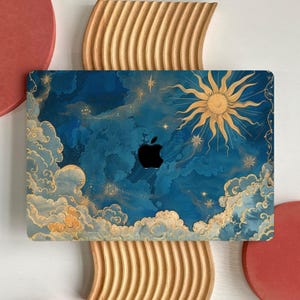 Op de afbeelding: Een laptop skin met een hemels ontwerp. Het heeft een zon, wolken en sterren in blauw, goud en wit. Het Apple-logo is zichtbaar. De skin is geplaatst op een houten decoratief stuk.