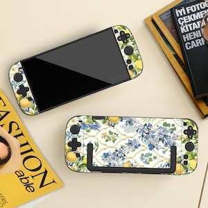 May include: Two Nintendo Switch consoles with floral and lemon-themed skins. The top console is open, displaying a black screen. The bottom console is closed, showcasing the patterned design. The design features yellow lemons, blue flowers, and green leaves.