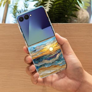May include: A clear phone case with a sunset design. The case features a vibrant stained-glass style image of a sunset over the ocean, with shades of yellow, blue, and gold. The phone is held in a hand.