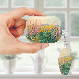 May include: A white and beige AirPod case and matching keychain featuring a colorful impressionist painting of a field of wildflowers. The case is held by a hand, showcasing the artwork's vibrant colors and design.