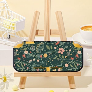 Pode incluir: Uma capa de consola de jogos portátil verde escuro com um padrão floral com flores rosa, amarelas e brancas e folhas verdes. A capa tem detalhes amarelos nas laterais e botões. A capa é exibida numa pequena tela de madeira.