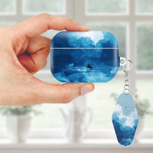 May include: A blue AirPod case with a matching keychain. The case and keychain feature a seascape design with a sailboat on the water under a cloudy sky. The case is held in a hand.